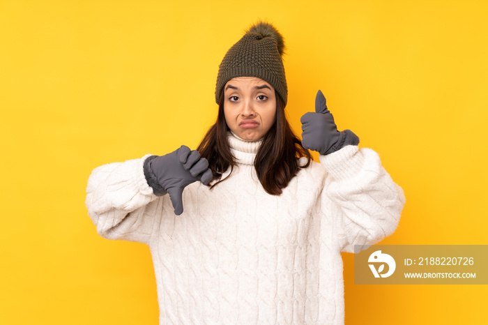 Young woman with winter hat over isolated yellow background making good-bad sign. Undecided between yes or not