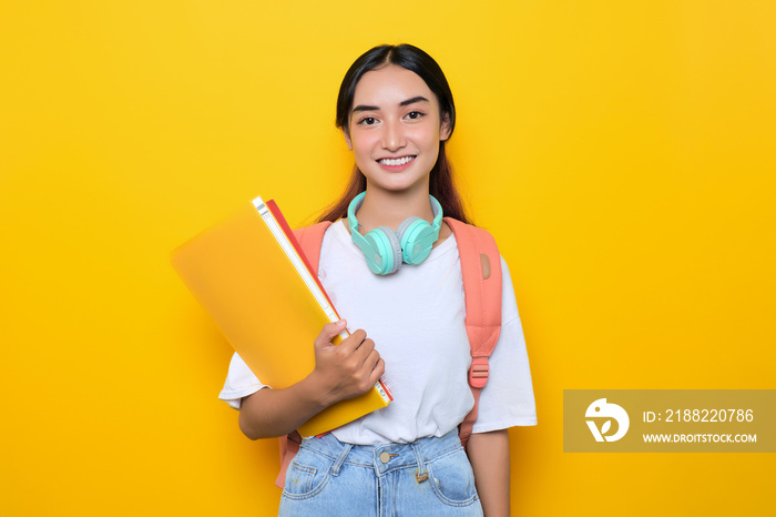 Cheerful pretty young student girl wearing backpack and headphones holding books isolated on yellow background