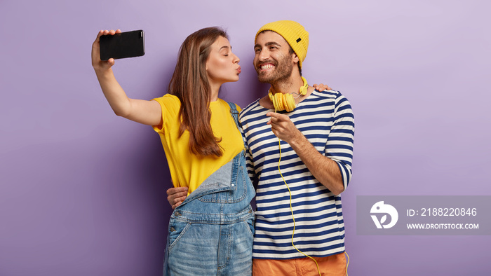 Loving couple stand together, embrace, kiss and take selfie, have perfect relationship. Affectionale girl in yellow t shirt and denim overalls, kisses boyfriend in cheek, holds cell phone, makes photo