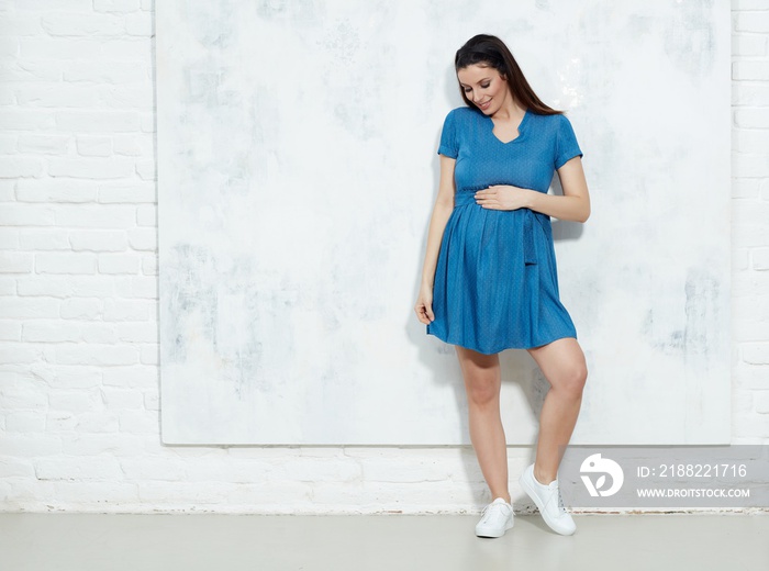 Fashion portrait of happy pregnant woman in blue summer dress against white wall with copyspace.