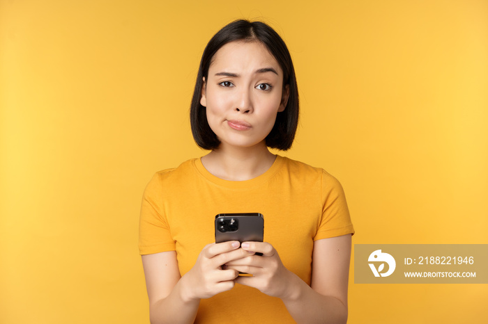 Skeptical asian woman holding smartphone, looking with doubt at camera, standing over yellow background