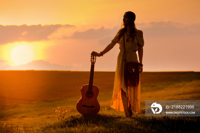 siluette of woman wearing a bohemian style holding a guitar on a field at warm light of sunset