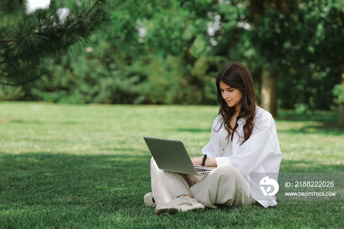 Beautiful Caucasian girl working with laptop computer sitting on green grass in summer park. Happy female outdoor working with technology, education and surfing the net, preparing for exams concept