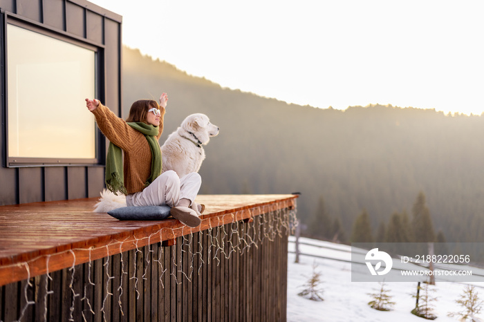 Woman sitting with dog on terrace of tiny house in the mountains enjoying beautiful sunrise landscape. Concept of small modern cabins for rest and escape to nature. Idea of traveling with dog