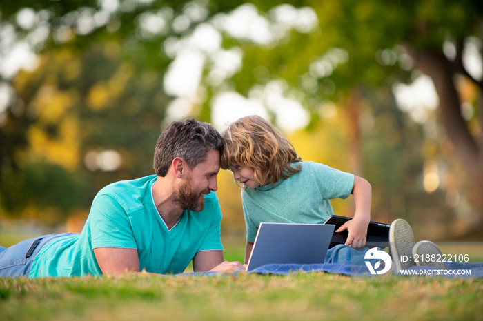 Weekend activity happy family lifestyle concept. Father with son use laptop. Dad and school boy child looking screen and tablet, watching video lesson, sitting on grass.