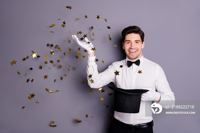 Portrait of positive cheerful illusionist hold cylinder hat make focus spell golden star confetti fly fall in air wear white shirt black bow isolated over grey color background