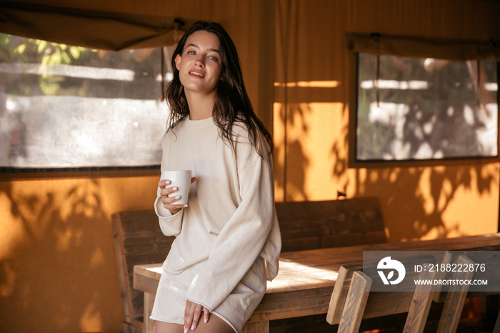 Pleasant young caucasian brunette woman posing looking at camera relaxing in country house. Lady with cup of tea wears casual clothes at home. Cozy spring concept