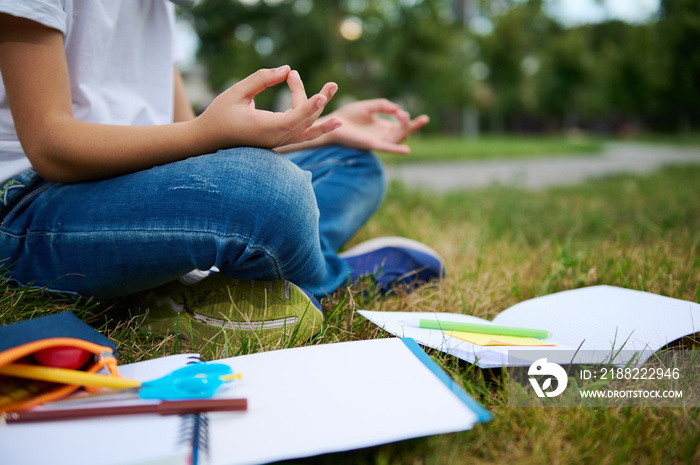 Cropped view of school child boy sitting in lotus position on green grass of city park and meditating . Workbooks school supplies lying on the grass. Concentration, recreation, mindfulness concepts