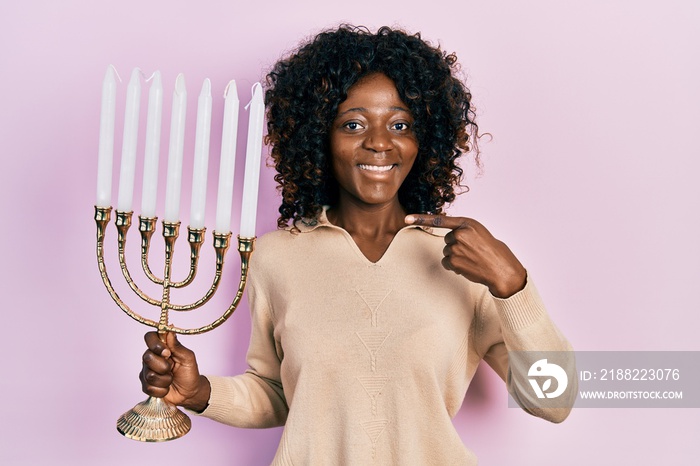 Young african american woman holding menorah hanukkah jewish candle smiling happy pointing with hand and finger