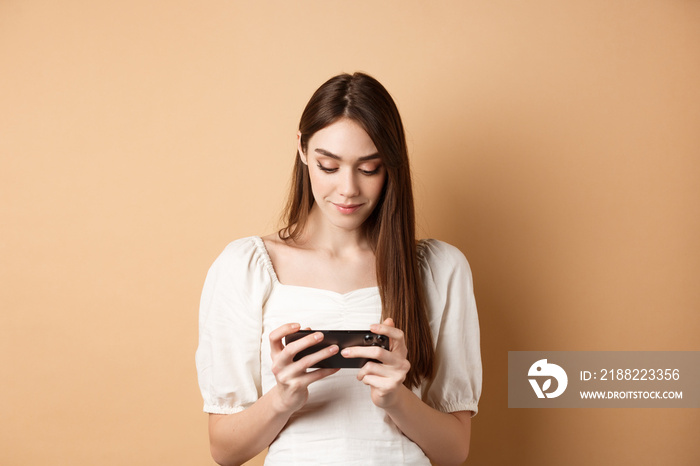 Pretty girl watching videos on smartphone, holding mobile phone horizontally and looking at screen, standing on beige background