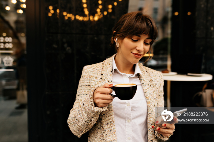 charming pretty european woman with dark hair wearing light jacket and blouse drinking coffee and using smartphone. Happy excited dark-haired woman laughing and walking around city