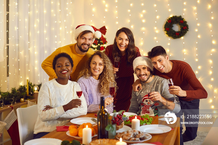 Happy diverse millennial friends at Christmas dinner at home. Group portrait of beautiful mixed race people holding wine glasses and looking at camera at festive table in cozy living room interior