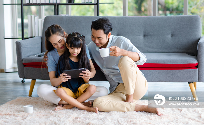 Millennial Asian happy family father drinking hot coffee while mother and dauthger sitting on cozy carpet floor smiling helping playing streaming remote game online on television in living room home