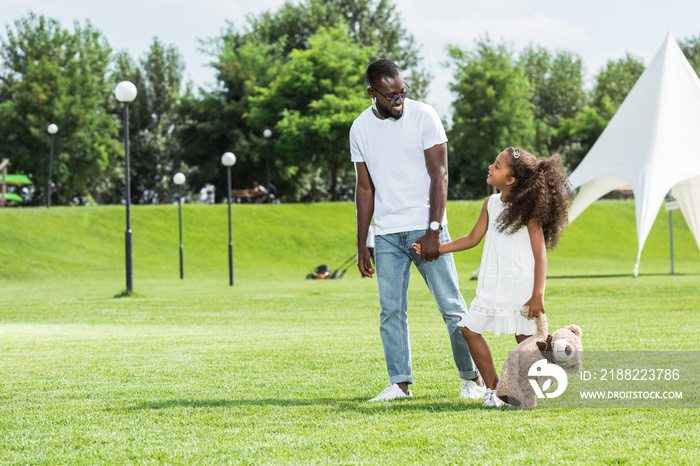 african american father and daughter with teddy bear holding hands and walking in park