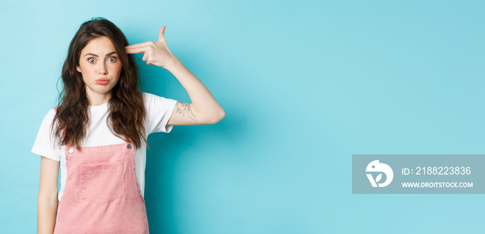 Image of tired and shocked young woman pointing finger gun at head, blowing her mind, staring bothered at camera, standing against blue background