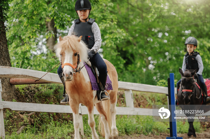 Children with helmets and protective vests on riding pony horses at sunny day on ranch.