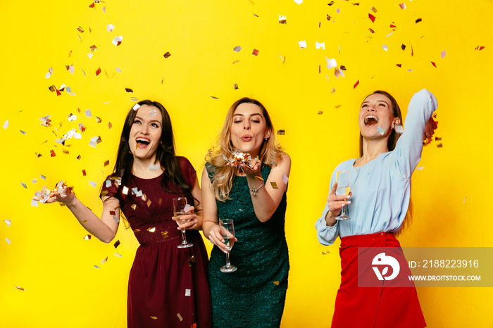 Happy March 8. Cheerful beautiful girls playing with confetti, blowing, having fun together while celebrating the women holiday, standing over yellow background. Dressed in colorful clothes.