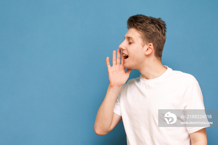 Portrait of a positive young man speaks and looks aside on a blue background. Smiling guy in a white T-shirt shouts an empty space for copyspace. Isolated