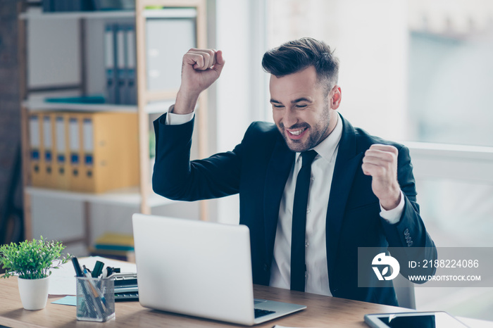 Portrait of young, smiling, attractive, confident finansist in tuxedo, blue jacket, triumphant  having his arms with fists raised, celebrating completed work, sitting at his desk top in work station