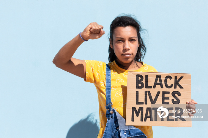 Portrait of Latin American woman with serious attitude and raised fist. She is isolated on blue background holding a banner with slogan Black Lives Matter. Space for text.