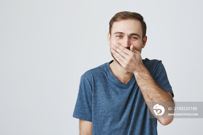 Positive human expressions concept. Bearded caucasian male in blue t-shirt laughs at good joke, hides his smile behind his hand. Laughing man posing against gray studio wall.