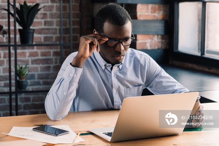 Serious african american businessman holding his eyeglasses, working with laptop in modern office