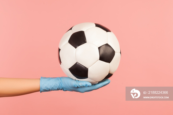 Profile side view closeup of human hand in blue surgical gloves holding football or soccer ball. indoor, studio shot, isolated on pink background.