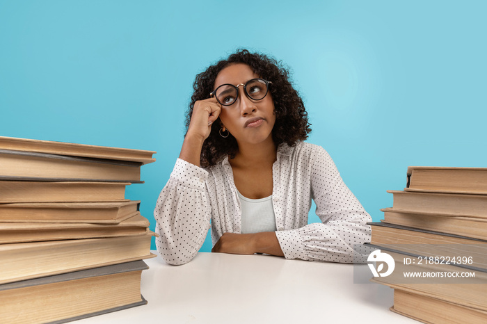 Young black female student in glasses sitting at desk with piles of books, daydreaming, feeling bored during lesson