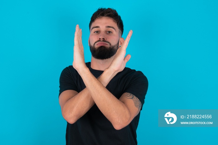 young bearded hispanic man wearing black T-shirt over blue background Rejection expression crossing arms doing negative sign, angry face