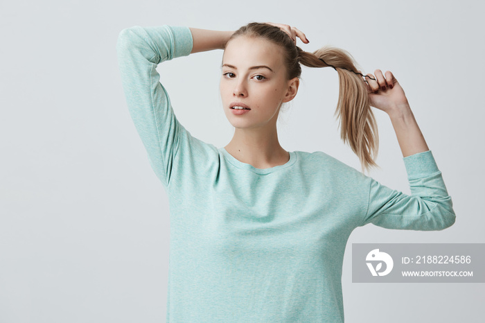 Good-looking young female with charming dark eyes and blonde hair in pony tail wearing light blue sweater, looking at camera with appeal, posing indoors. Beauty and youth concept