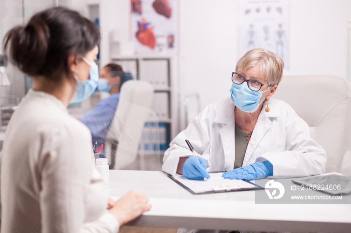 Doctor wearing protection mask against covid taking notes during consultation with patient in medical clinic. Nurse wearing blue uniform while working on computer.