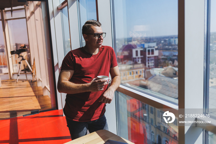 A young man in a red t-shirt, glasses for vision correction on his face. He stands at the window with a cup of coffee and looks at the city.