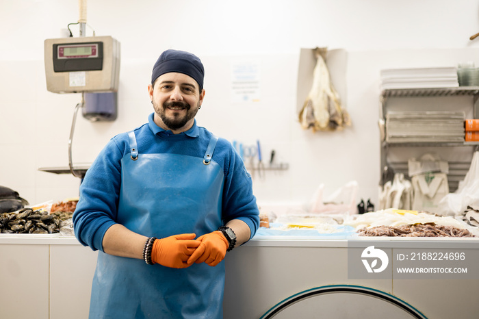 A middle-aged fishmonger poses in front of the product counter leaning while looking at the camera, selling food, small and medium business concept.