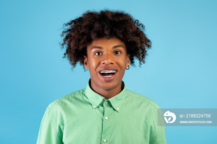 Excited african american teen guy with braces looking and smiling at camera, posing over blue studio background