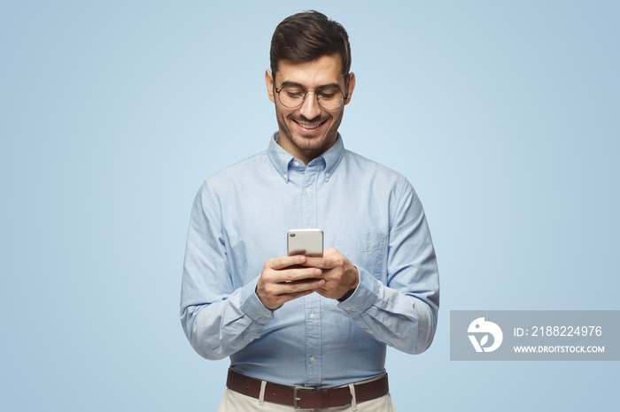 Portrait of cheerful businessman in round glasses typing sms with phone on blue background