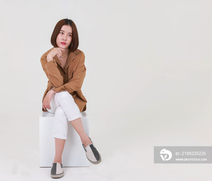 Portrait studio cutout shot of Asian young pretty short hair female model in long brown coat jacket casual shoes sitting crossed legs smiling look at camera on box chair posing on white background