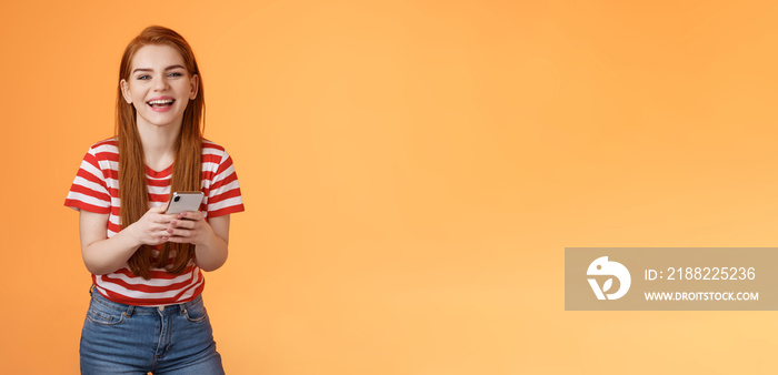 Carefree attractive woman red long hair laughing amused, having fun, chatting friend, hold smartphone, look camera sincere rejoice smile, texting joke, found funny meme internet, orange background