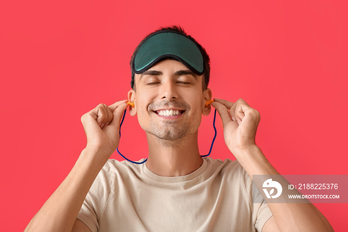 Smiling young man with sleeping mask putting ear plugs on red background