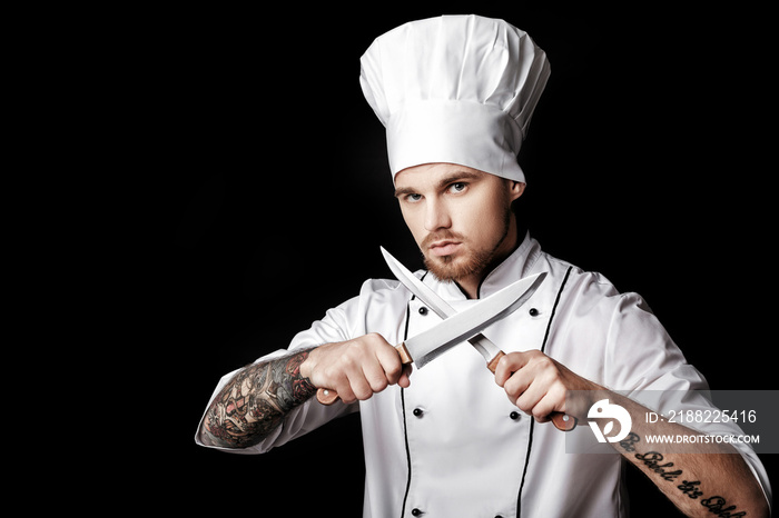 Young bearded man chef In white uniform holds Two knives on  black background