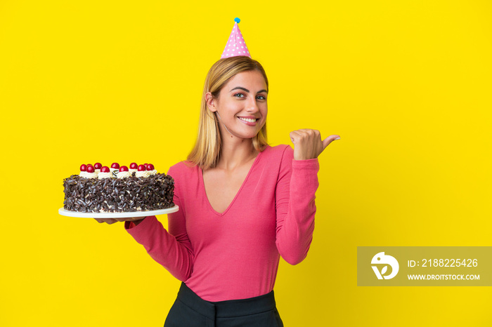 Blonde Uruguayan girl holding birthday cake isolated on yellow background pointing to the side to present a product