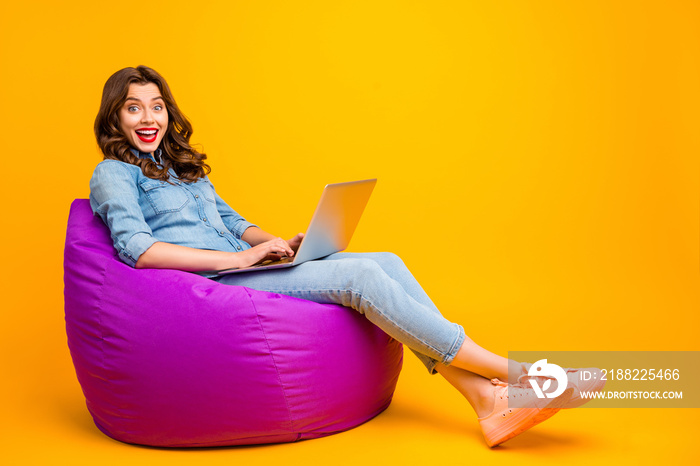 Full length body size turned photo of cheerful cute positive nice pretty girl browsing through computer with excited facial expression and legs crossed isolated over yellow vibrant color background