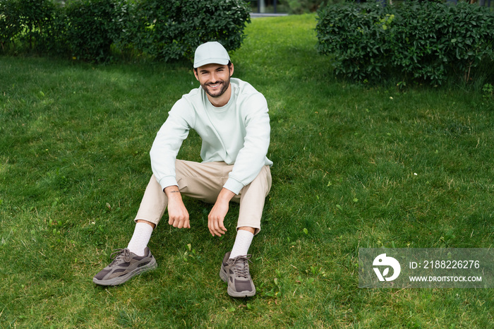 full length of bearded man in cap smiling while sitting on green lawn