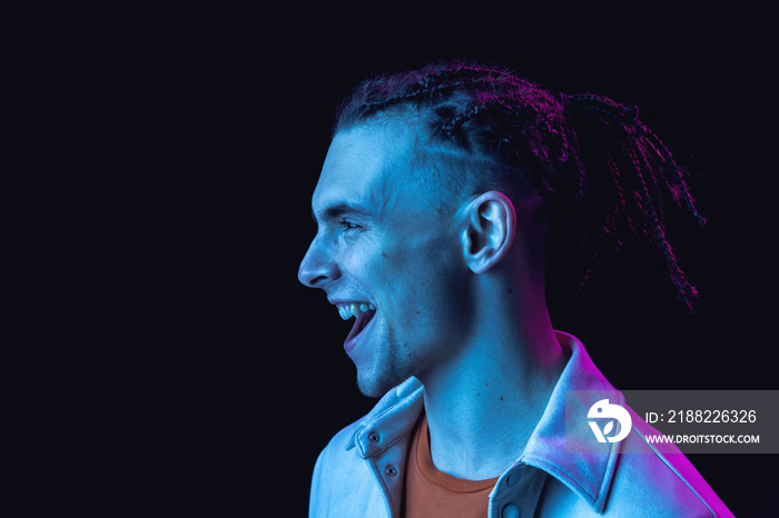 Caucasian young man’s portrait on dark studio background in neon. Concept of human emotions, facial expression.