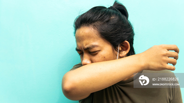 A portrait of a Malay man coughing and sneezing to his elbow with closing both eyes on isolated blue background