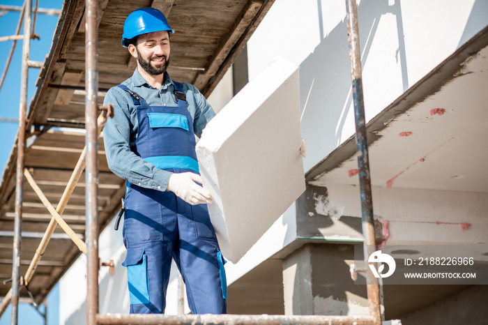 Builder warming a building facade with foam panels standing on the scaffoldings on the construction site