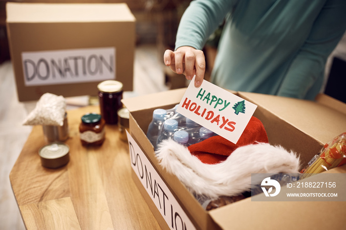 Close-up of woman donating food for charity and adding greeting card with ’Happy Holidays’ message into a box.