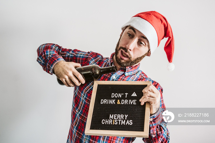 .Young man disguised as Santa Claus, drinking wine clearly drunk with a serious message on his blackboard  Don’t drink and drive, Merry Christmas . Serious message.