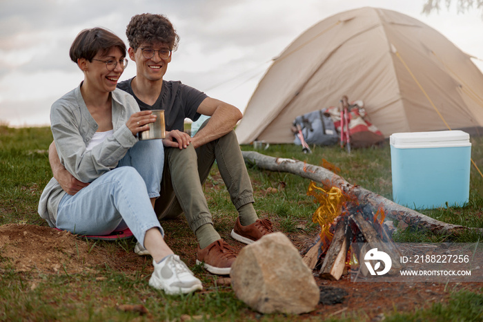 Man and woman drinking tea or coffee at fire in camp stop. Enjoying summer day.