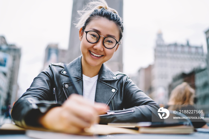 Portrait of happy Chinese tourist sitting in street cafe and smiling at camera enjoying leisure time on vacations, cheerful Japanese hipster girl in trendy spectacles for provide eyes correction