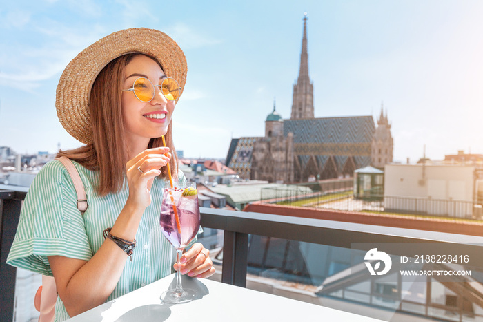 A cheerful girl in a hat drinks an alcoholic cocktail on the terrace of the bar with a great view of the city of Vienna with St. Stephen’s Cathedral